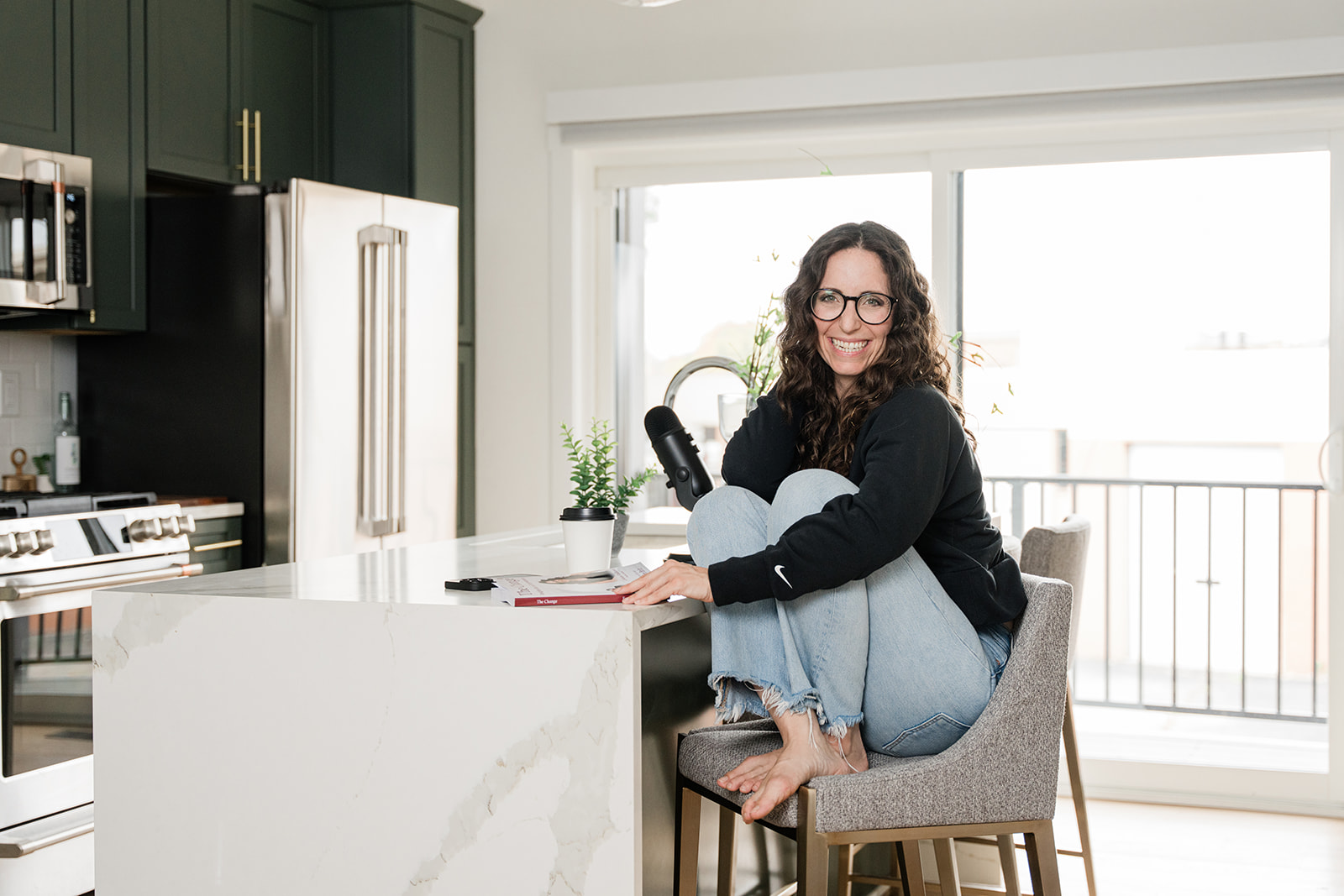 Lindsaya casual on a stool with her mic and a copy of The Change series.
